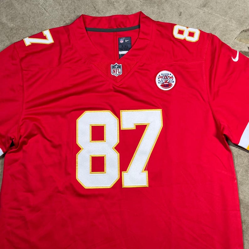 May include: A red NFL jersey with white and gold accents. The jersey features the number 87 in large white block letters with gold trim on the front. The number 8 and 7 are also visible on the shoulders. The NFL and Nike logos are also visible.