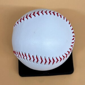 May include: A white baseball with red stitching. The ball is sitting on a black surface. The baseball is a classic sports item.