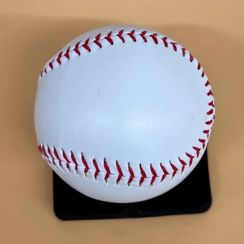 May include: A white baseball with red stitching. The ball is sitting on a black surface. The baseball is a classic sports item.