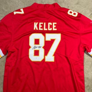 May include: Red football jersey with the name "KELCE" and the number "87" in white and gold. The number "87" is on the front and the back of the jersey. The number "7" is on the left shoulder and the number "8" is on the right shoulder.