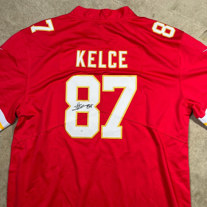 May include: Red football jersey with the name "KELCE" and the number "87" in white and gold. The number "87" is on the front and the back of the jersey. The number "7" is on the left shoulder and the number "8" is on the right shoulder.