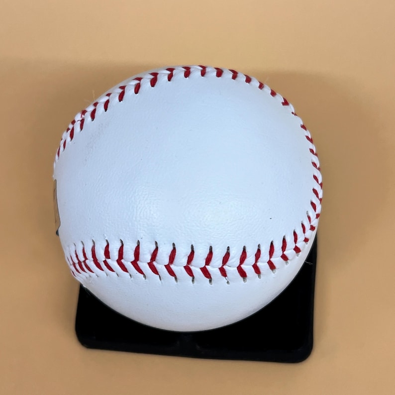 May include: A white baseball with red stitching. The ball is resting on a black surface. The baseball is a classic sports item.