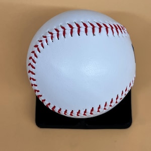 May include: A white baseball with red stitching, resting on a black surface. The baseball is a classic design, with visible seams and a smooth, clean surface. The stitching is a vibrant red, contrasting against the white leather.