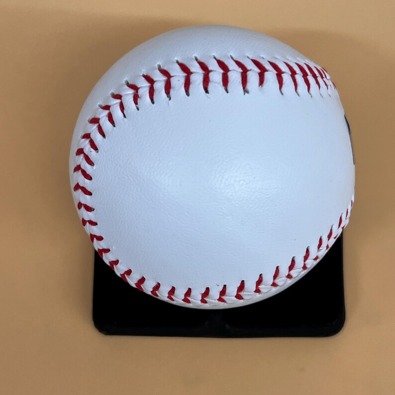 May include: A white baseball with red stitching, resting on a black surface. The baseball is a classic design, with visible seams and a smooth, clean surface. The stitching is a vibrant red, contrasting against the white leather.