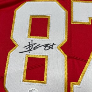 May include: Close-up of a red jersey with a large white number 84 outlined in gold. The number has a red interior. A black signature is across the number. The jersey has a VS Autograph Authentication sticker.