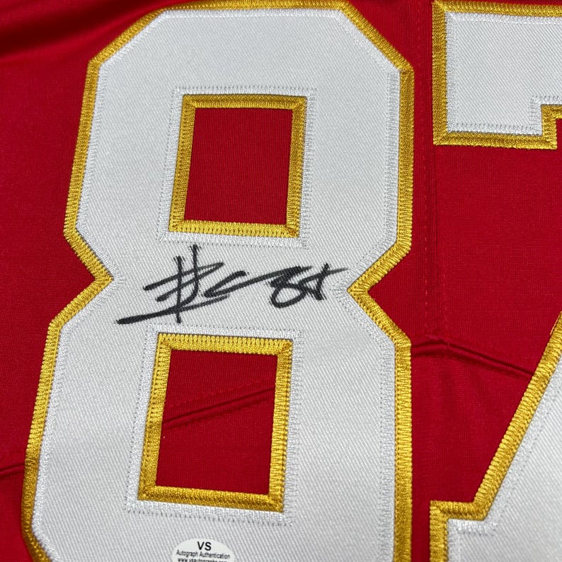 May include: Close-up of a red jersey with a large white number 84 outlined in gold. The number has a red interior. A black signature is across the number. The jersey has a VS Autograph Authentication sticker.