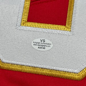 May include: Close-up of a red fabric with a white and gold embroidered patch. The patch has a white border with gold stitching and a red center. A white sticker with black text is affixed to the fabric, reading "VS Autograph Authentication."