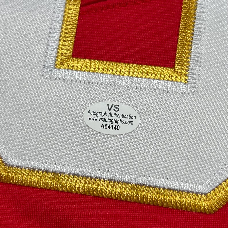 May include: Close-up of a red fabric with a white and gold embroidered patch. The patch has a white border with gold stitching and a red center. A white sticker with black text is affixed to the fabric, reading "VS Autograph Authentication."