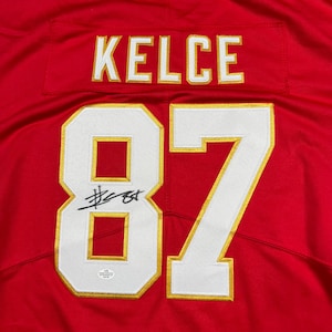 May include: A red jersey with the name "KELCE" in white and gold lettering across the top. The number "87" is prominently displayed in white with gold trim. A signature is visible on the number 87.