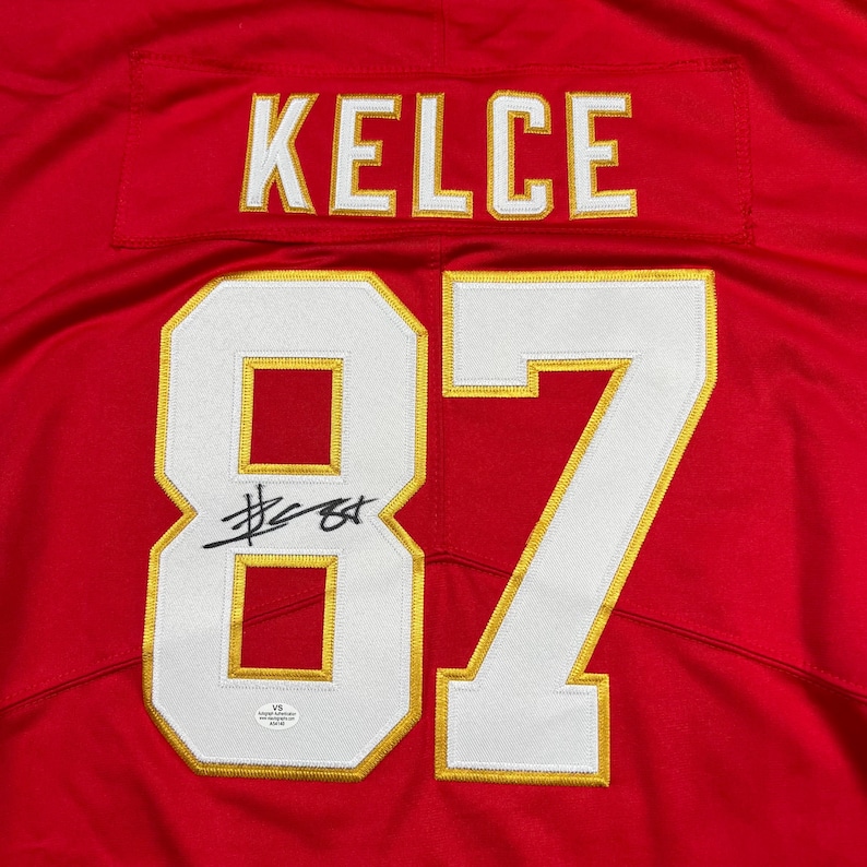 May include: A red jersey with the name "KELCE" in white and gold lettering across the top. The number "87" is prominently displayed in white with gold trim. A signature is visible on the number 87.
