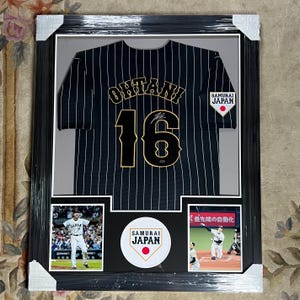 Pode incluir: Camisola de beisebol a preto e branco emoldurada com o nome "Ohtani" e o número "16" em letras douradas. A camisola é assinada e apresenta o logótipo "Samurai Japan". Inclui duas fotos de ação e um logótipo.