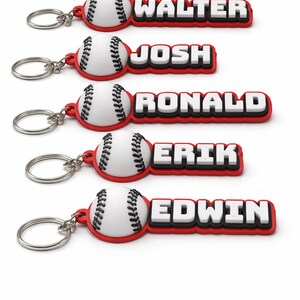 Könnte beinhalten: Fünf Schlüsselanhänger im Baseball-Design mit einem weißen Ball mit schwarzen Nähten, der an einem roten rechteckigen Anhänger befestigt ist. Jeder Anhänger zeigt einen Namen in weißen Blockbuchstaben mit schwarzem Rand: WALTER, JOSH, RONALD, ERIK und EDWIN.