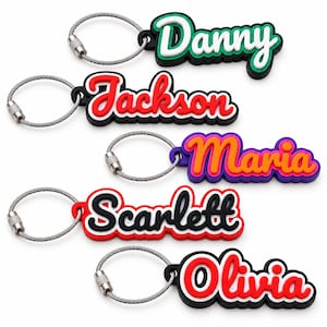 May include: Five custom name keychains featuring flexible, multi-layered rubberized lettering in various colors including green, red, purple, black, and white. Each keychain is attached to a circular, silver-toned braided metal wire cable loop.