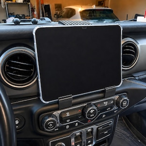 Könnte beinhalten: Ein schwarzes Tablet, das auf dem Armaturenbrett eines Fahrzeugs über dem Klimabedienteil montiert ist. Das Tablet wird von einer dunkelgrauen Kunststoffhalterung gehalten, die an der Mittelkonsole befestigt ist und eine transparente Schutzhülle aufweist.