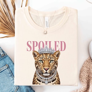 May include: Cream-colored t-shirt featuring a leopard wearing a crown and a pearl necklace. The word "SPOILED" is printed above the leopard in pink letters. A silver necklace is visible. The shirt is folded and placed on a white surface.