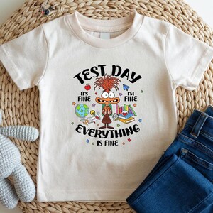 May include: A cream-colored t-shirt with a colorful graphic that reads "TEST DAY" and "EVERYTHING IS FINE." The graphic features a cartoon character, an apple, books, and a globe. Blue denim jeans are visible.