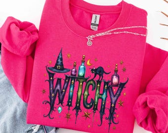 May include: A bright pink crewneck sweatshirt featuring a graphic print with the word WITCHY in large, stylized, multi-colored letters. The design includes a black witch hat, a black cat, small potion bottles, stars, and a crescent moon.