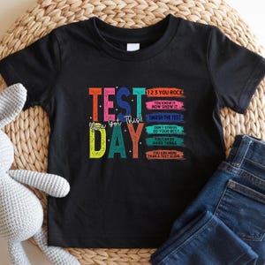 May include: Black t-shirt with the words "TEST DAY" in colorful block letters. The shirt also has the text "You Got This!" and phrases like "1 2 3 You Rock" and "Smash the Test".