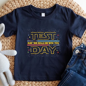 May include: Navy blue short-sleeve t-shirt with the text "TEST DAY" in yellow, and "MAY THE SKILLS BE WITH YOU" in white. The design includes illustrations of lightsabers in red and blue. The shirt is paired with blue jeans.