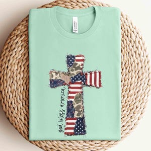 May include: A light mint green t-shirt featuring a central cross graphic composed of patchwork patterns including the American flag, camouflage, and blue star prints. The text "god bless america" is printed in black script to the left of the cross.