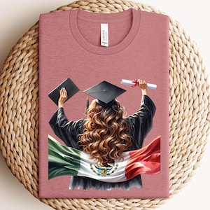 May include: Dusty rose t-shirt featuring a graduation design. The graphic shows a graduate in a cap and gown holding a diploma and a book, with the Mexican flag draped behind them. The word "Canvas" is printed on the tag.