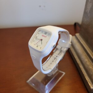 K&ouml;nnte beinhalten: Eine wei&szlig;e Rip Curl-Uhr mit quadratischem Zifferblatt und wei&szlig;em Armband. Die Uhr hat einen roten Sekundenzeiger und den Markennamen "ripcurl" in Rot. Die Uhr wird auf einem transparenten Acryl-St&auml;nder pr&auml;sentiert.