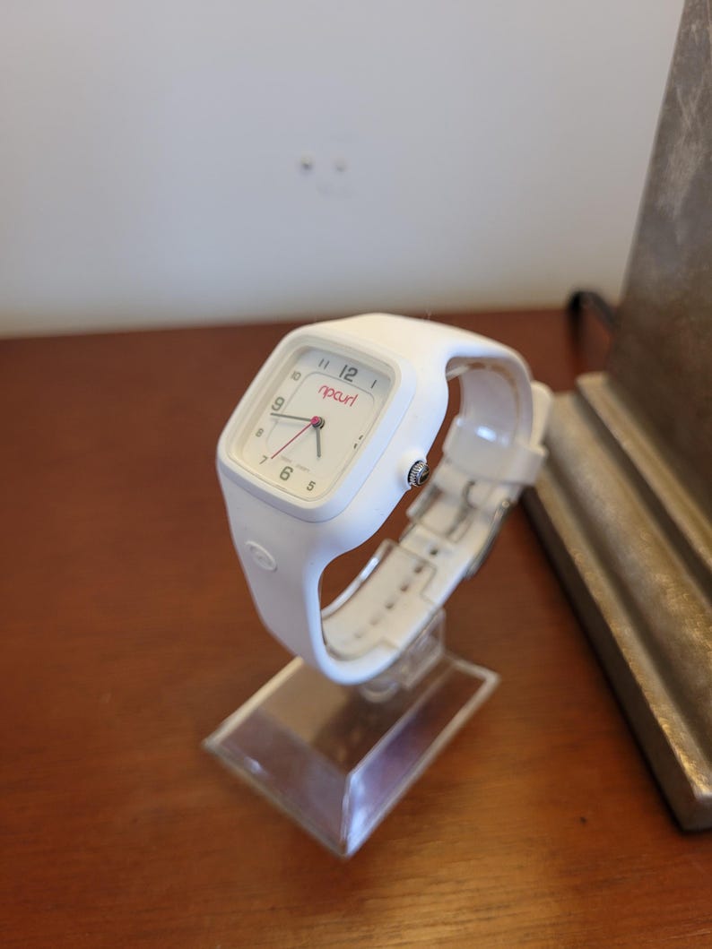 K&ouml;nnte beinhalten: Eine wei&szlig;e Rip Curl-Uhr mit quadratischem Zifferblatt und wei&szlig;em Armband. Die Uhr hat einen roten Sekundenzeiger und den Markennamen "ripcurl" in Rot. Die Uhr wird auf einem transparenten Acryl-St&auml;nder pr&auml;sentiert.