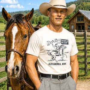 May include: A cream-colored short-sleeve t-shirt featuring a black graphic print of a person riding a horse. The text reads Darling Ranch Resort and Alexandria, Minn. The shirt is paired with grey trousers and a black belt.