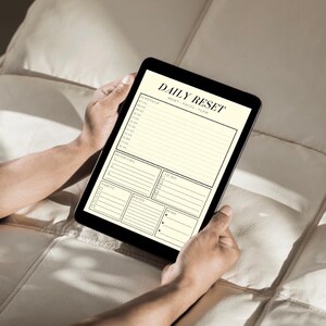 May include: A black tablet displaying a "DAILY RESET" planner with sections for schedule, priorities, to-do list, habits, notes, and wins. The tablet is held by hands against a neutral-toned background.