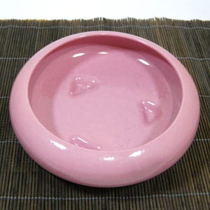 May include: A round, pale pink ceramic bowl with a glossy finish. The bowl has a wide rim and three raised, heart-shaped details inside. The bowl is set on a brown, woven mat.