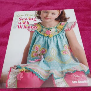 May include: A softcover book titled More Sewing with Whimsy by Kari Mecca, published by Martha Pullen. The cover features a child wearing a light blue dress with white polka dots, floral applique, and pink trim, paired with solid pink pants.