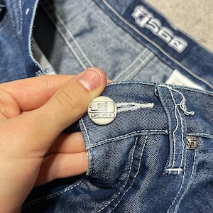 Puede incluir: Primer plano de unos vaqueros azules con costuras blancas. Los vaqueros tienen un bot&oacute;n plateado con el texto "S60" y una etiqueta plateada a juego. Los vaqueros est&aacute;n doblados, mostrando el bolsillo y la etiqueta de la marca.