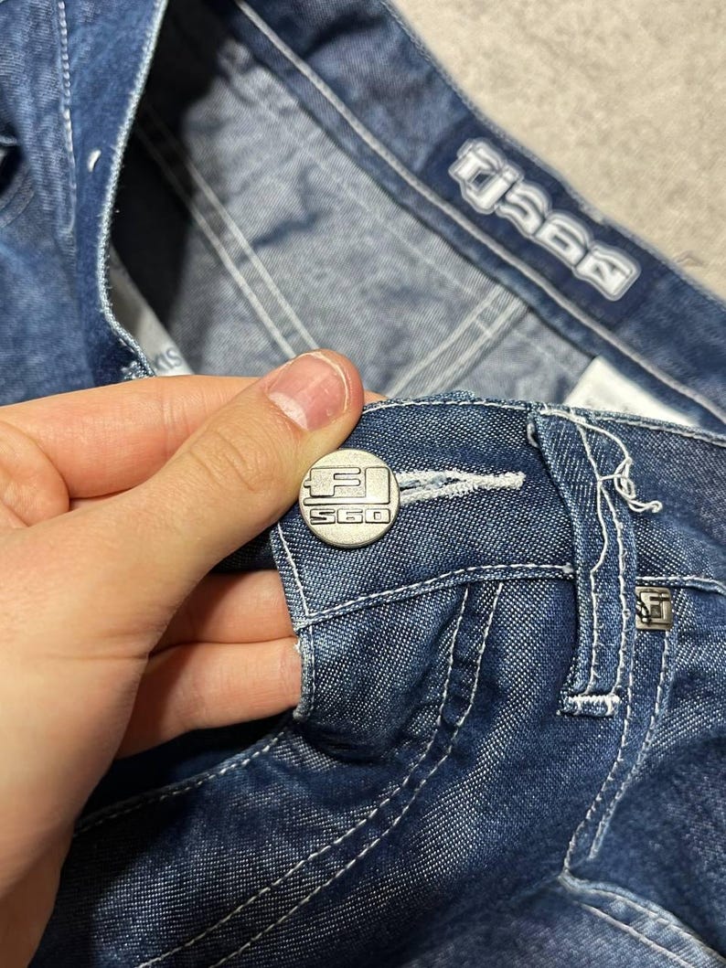 Puede incluir: Primer plano de unos vaqueros azules con costuras blancas. Los vaqueros tienen un bot&oacute;n plateado con el texto "S60" y una etiqueta plateada a juego. Los vaqueros est&aacute;n doblados, mostrando el bolsillo y la etiqueta de la marca.