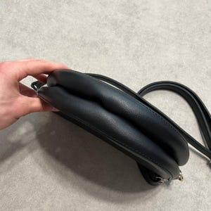 May include: A black, rounded crossbody bag with a long strap. The bag has a smooth, leather-like texture and a small, gold-colored clasp. The bag is held by a hand.