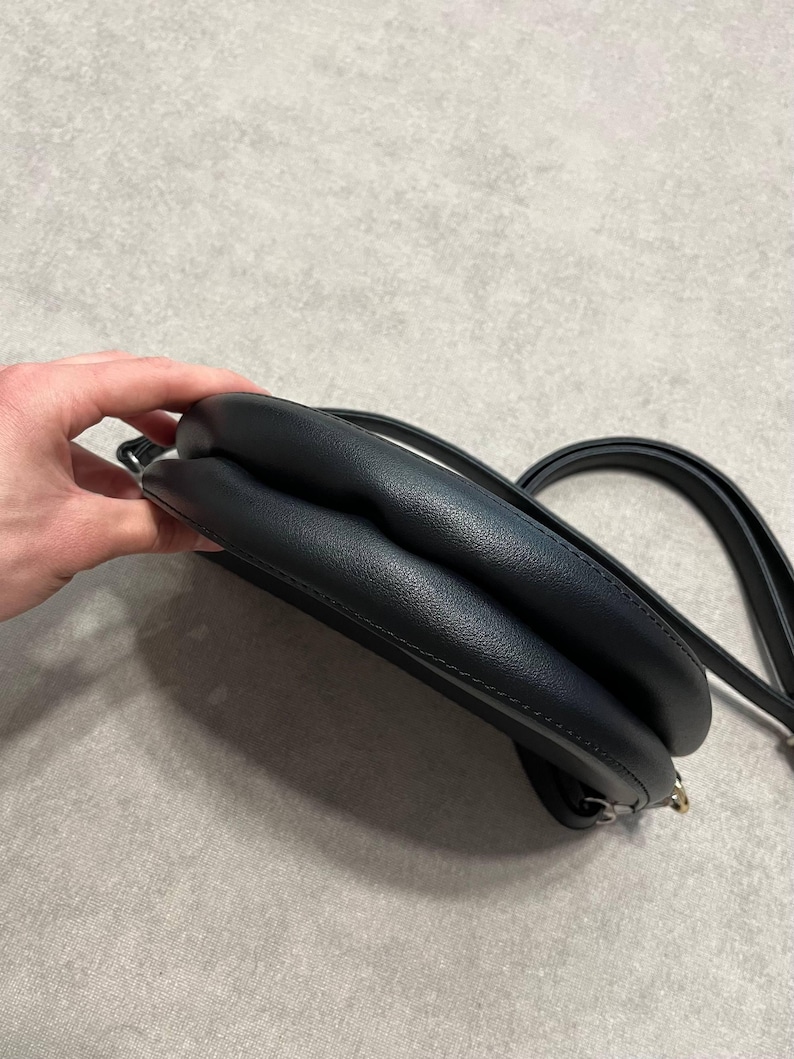 May include: A black, rounded crossbody bag with a long strap. The bag has a smooth, leather-like texture and a small, gold-colored clasp. The bag is held by a hand.