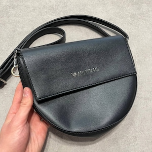 May include: Black crossbody bag with a semi-circular design. The bag features a flap closure and a long, adjustable strap. The word "VALENTINO" is embossed on the front. The bag is made of a smooth, leather-like material.