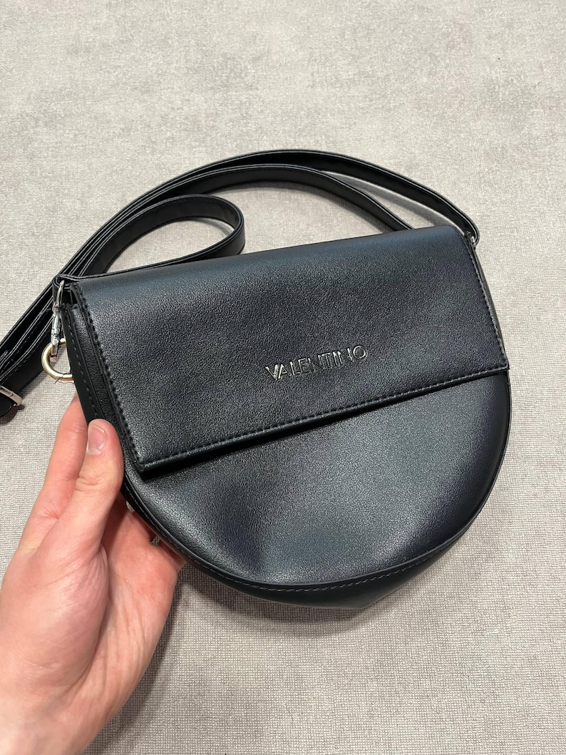 May include: Black crossbody bag with a semi-circular design. The bag features a flap closure and a long, adjustable strap. The word "VALENTINO" is embossed on the front. The bag is made of a smooth, leather-like material.