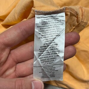 May include: Close-up of a peach-colored garment with a care label. The label features multiple lines of text in various languages, including English, French, and German, with warnings about fire and heat.