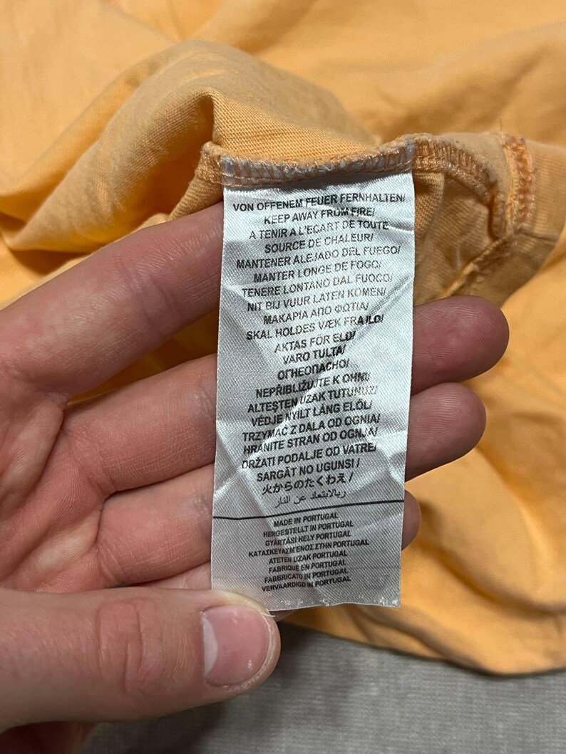 May include: Close-up of a peach-colored garment with a care label. The label features multiple lines of text in various languages, including English, French, and German, with warnings about fire and heat.
