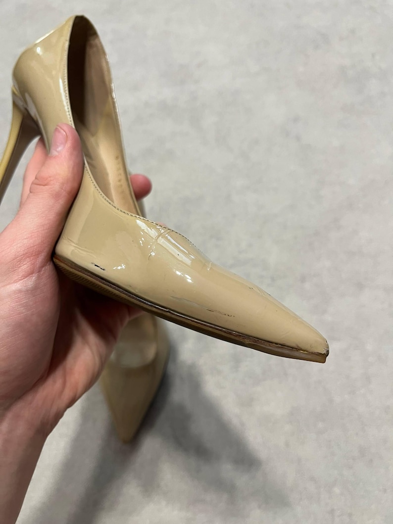 Puede incluir: Un par de zapatos de tac&oacute;n alto de charol beige con punta. Los tacones de aguja son delgados y los zapatos tienen un acabado brillante. Los zapatos est&aacute;n sostenidos por una mano.