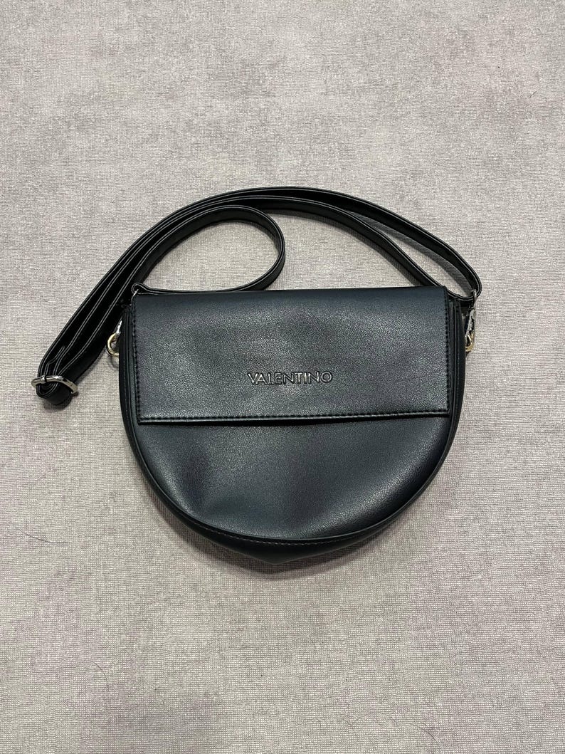 May include: Black crossbody bag with a curved bottom and a flap closure. The bag has a long, adjustable strap and the brand name "VALENTINO" is embossed on the front. The bag is made of a smooth, matte material.