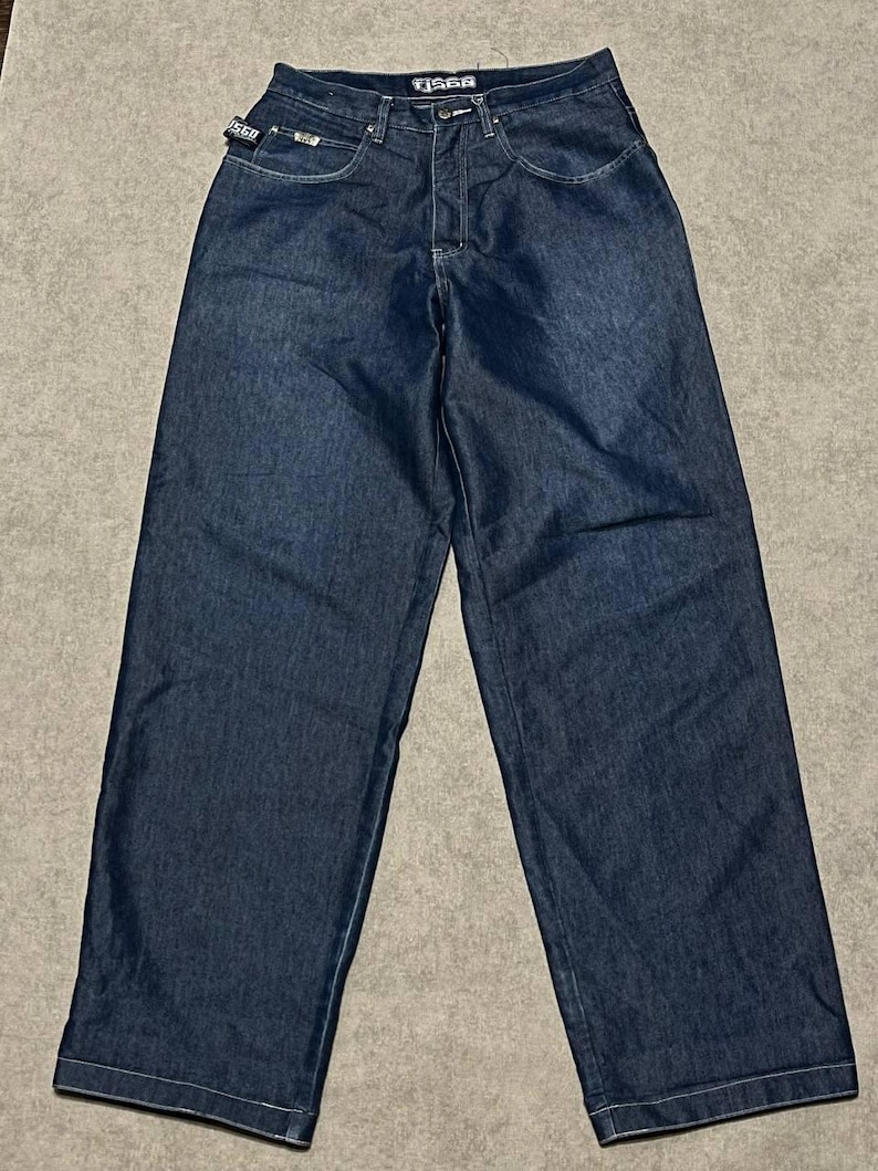 Puede incluir: Vaqueros de mezclilla azul oscuro con dise&ntilde;o de pernera ancha. Los vaqueros tienen un estilo cl&aacute;sico de cinco bolsillos, cierre de bot&oacute;n y cremallera, y el nombre de la marca "FIS6R" es visible en la cintura. Los vaqueros est&aacute;n planos sobre una superficie neutra.