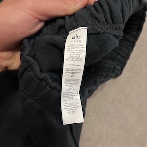 May include: Dark charcoal sweatpants featuring an elasticized waistband and an interior care label with the brand name Alo, size S/P/CH, and material composition of 65% cotton and 35% polyester, manufactured in Vietnam with a soft fleece lining.