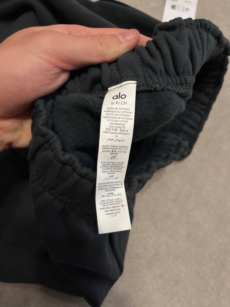 May include: Dark charcoal sweatpants featuring an elasticized waistband and an interior care label with the brand name Alo, size S/P/CH, and material composition of 65% cotton and 35% polyester, manufactured in Vietnam with a soft fleece lining.