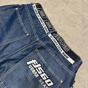 Puede incluir: Primer plano de unos vaqueros azules. Los vaqueros presentan texto blanco y negro "FJ 560" y "Urban Gear" en los bolsillos traseros y la cintura. Los vaqueros tienen un dise&ntilde;o cl&aacute;sico de cinco bolsillos y corte recto.