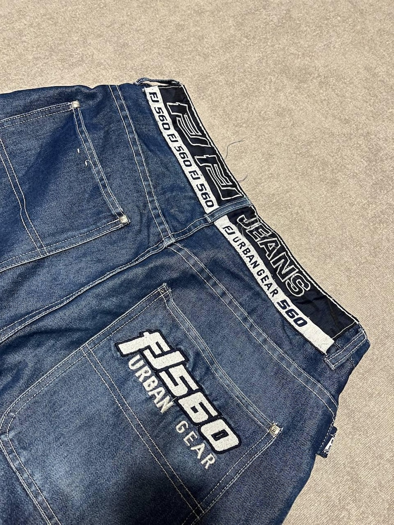 Puede incluir: Primer plano de unos vaqueros azules. Los vaqueros presentan texto blanco y negro "FJ 560" y "Urban Gear" en los bolsillos traseros y la cintura. Los vaqueros tienen un dise&ntilde;o cl&aacute;sico de cinco bolsillos y corte recto.