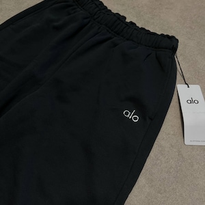 May include: Black sweatpants featuring an elasticized waistband and a white embroidered logo on the upper left side. A white rectangular paper tag is attached to the waistband with a black string, displaying the text alo and ALOYOGA.COM.