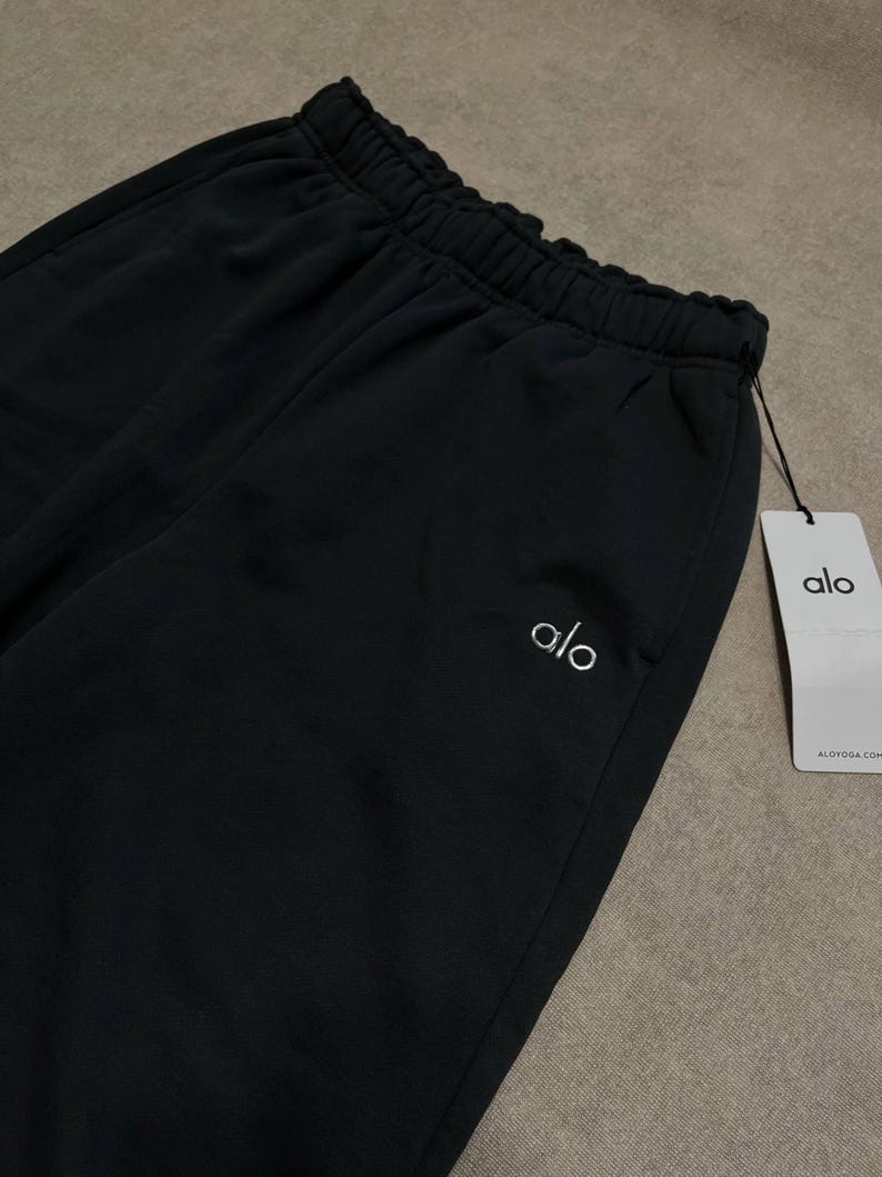 May include: Black sweatpants featuring an elasticized waistband and a white embroidered logo on the upper left side. A white rectangular paper tag is attached to the waistband with a black string, displaying the text alo and ALOYOGA.COM.