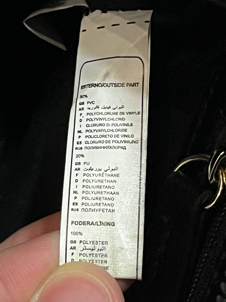 May include: A close-up of a tag with text in multiple languages, including English, French, and Spanish. The tag lists the materials used in a product, such as PVC, polyurethane, and polyester. The tag is white with black text.