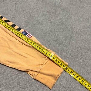 May include: A long-sleeved, peach-colored shirt sleeve with a black and white stripe design and a small American flag patch. A yellow measuring tape is laid across the sleeve, showing measurements in inches.
